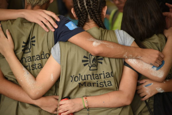 Jovens de costas abraçadas em um círculo com o uso de coletes escrito “Voluntariado Marista.” 